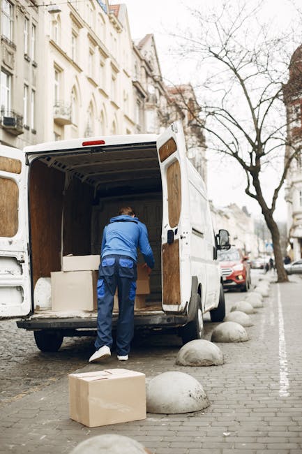 A professional mover from Removal Companies Finchley is loading a large cardboard box into the rear of a white moving van parked on a cobblestone street. The van's back doors are open, revealing an interior with wooden paneling and stacked furniture wrapped in protective materials. The mover, dressed in a blue jacket and dark trousers, is bent over, carefully placing the box inside the vehicle. Several other boxes, some wrapped in plastic or packing paper, are positioned on the sidewalk nearby, ready for transportation. The scene is set in an urban residential area with multi-story buildings featuring classic architectural details, leafless trees lining the pavement, and a few parked cars, including a red vehicle further down the street. This image illustrates the process of home relocation, with professional furniture transport and loading, as part of a comprehensive removal service offered by [COMPANY_NAME], emphasizing careful packing and efficient moving logistics.