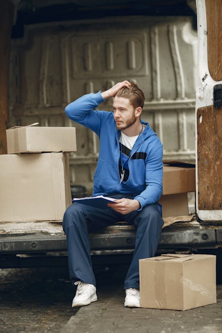 A young man with light brown hair and a beard, wearing a blue hoodie and dark trousers, sits on the edge of an open moving van. He is holding a clipboard in his left hand and scratching his head with his right, appearing to be reviewing a packing list or inventory. Surrounding him are several cardboard boxes, some stacked inside the van and others placed on the ground nearby, indicating a home relocation process. The interior of the van has a plain, industrial appearance with a metallic wall and a wooden panel on the side. The scene takes place outdoors on a paved surface, suggesting the loading or unloading phase of furniture transport and moving logistics as part of the services offered by Removal Companies Finchley. The lighting is natural, highlighting the details of the boxes, the man's expression, and the moving equipment involved in the packing and moving process.