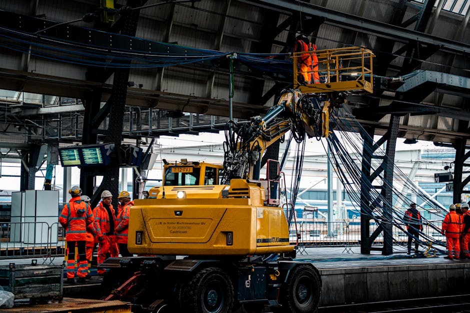 Inside Finchley Central station, a large yellow telescopic boom lift with an extended arm is positioned beneath an overhead structure, allowing a worker in a high-visibility orange vest and helmet to perform maintenance or inspection on the station's ceiling or lighting fixtures. Several other workers, also in high-visibility orange clothing and safety helmets, stand on the adjoining platform, observing and assisting with the operation. The station's interior features metallic beams, panels, and digital display screens hanging from the ceiling, providing information for travelers. In the background, part of the station's platform and interior architecture are visible, with natural daylight filtering through open sections near the roof. This scene reflects a professional maintenance or inspection process, emphasizing safe working practices and equipment used in station management, aligned with the services offered by Removal Companies Finchley for home or commercial relocations involving station access or logistical challenge areas.