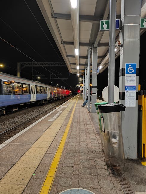 Nighttime view of Finchley Central station platform showing a waiting area with a covered canopy supported by metal poles. On the platform, there is a green and black trash bin, a blue and white designated accessible parking sign attached to a pole, and a large silver trash receptacle. To the left, a modern train with blue and white carriages is stationary on the tracks, with visible windows illuminated from inside. The platform surface is paved with concrete slabs and tactile paving strips near the edge for safety. Overhead, fluorescent lighting lines the canopy, providing visibility despite the dark night sky. The scene represents a typical setting for a home relocation or moving logistics service, where furniture or boxes might be transported to or from the station as part of a house removals process facilitated by Removal Companies Finchley.