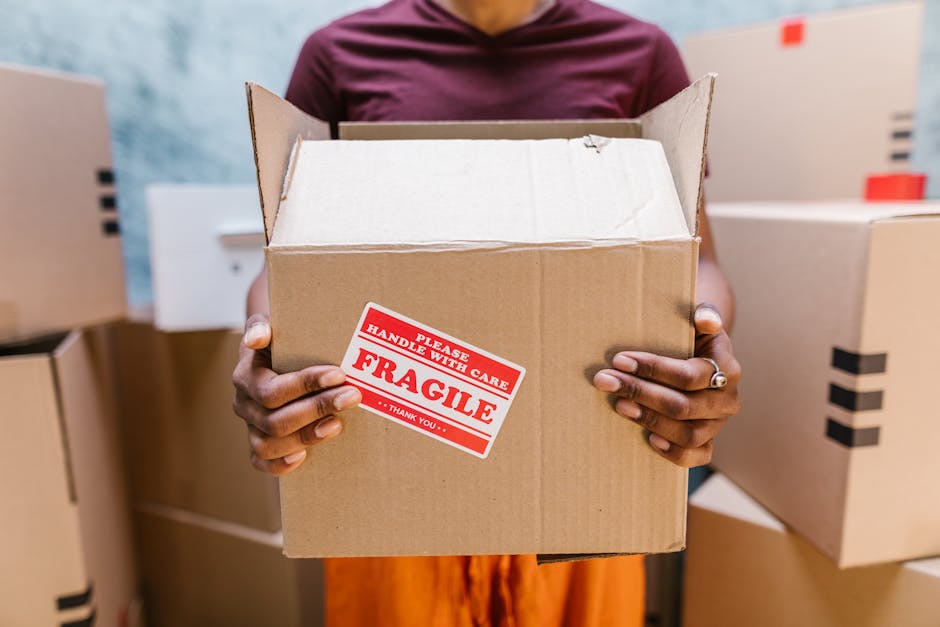 A person wearing a maroon shirt and orange pants is holding an open cardboard box marked with a red and white 'Fragile Handle with Care' sticker, situated inside a room with a light blue wall. Several closed cardboard boxes are stacked on shelves and the floor around them, some with black tape sealing the lids. The person’s hands are visible grasping the box, with their face not shown. The scene reflects home relocation or moving preparations, emphasizing the careful handling of fragile items during packing and furniture transport. This setting aligns with the services offered by Removal Companies Finchley for house removals and moving logistics, illustrating the packing and loading process involved in residential moves.