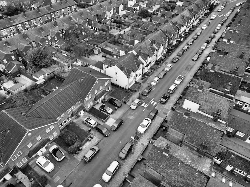 An aerial black-and-white photograph of a residential street showing a row of houses with pitched roofs, small backyards, and garden sheds, alongside parked cars lining both sides of the street. The image captures an area where home relocation or furniture transport may occur, with a focus on the narrow roadway and residential parking arrangements. Removal Companies Finchley are experienced in managing packing and loading processes, and the visible vehicles and street layout highlight typical urban moving logistics and parking considerations for house removals on North Finchley High Road. The scene includes an adjacent pavement, street lamps, and small front gardens, illustrating the typical environment where professional moving services facilitate efficient logistics during a house move or furniture transport in this urban neighborhood.