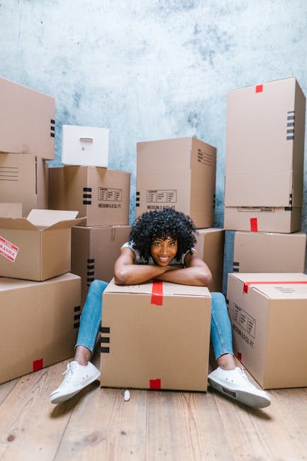 A young woman with curly black hair, wearing a grey T-shirt, blue jeans, and white sneakers, is sitting on a wooden floor surrounded by numerous cardboard moving boxes, some sealed with red packing tape and others partially open revealing packing materials inside. She is resting her chin and arms on a large box in front of her, smiling at the camera. The background features a textured light blue wall, and the scene appears to be inside a room prepared for a home relocation, with boxes stacked in various positions around her. The image captures the process of packing and preparing for a house move, often associated with professional removals services such as those offered by Removal Companies Finchley, supporting furniture transport and packing during a local move.