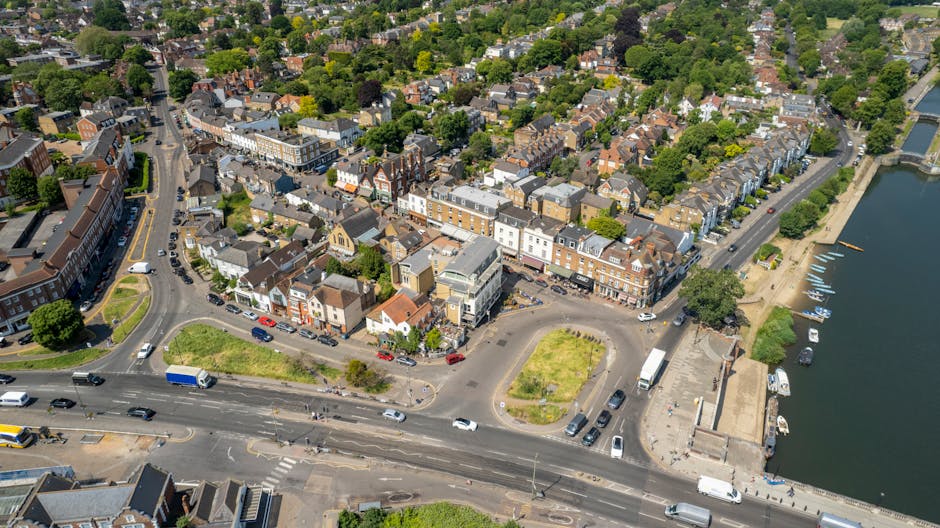 An aerial view of a riverside urban area showing a mix of residential and commercial buildings with rooftops in various shades of brown and grey, surrounded by trees and green spaces. At the bottom of the image, a busy road with multiple lanes is visible, with cars, vans, and a bus traveling in both directions. Near the water, there is a paved area with parked boats and a small dock, while a pathway runs along the riverbank. The scene depicts a typical town layout, with streets radiating from a central point and extending towards the river, illustrating the kind of environment in which house removals and furniture transport services, such as those provided by Removal Companies Finchley, may operate during home relocations in the area.